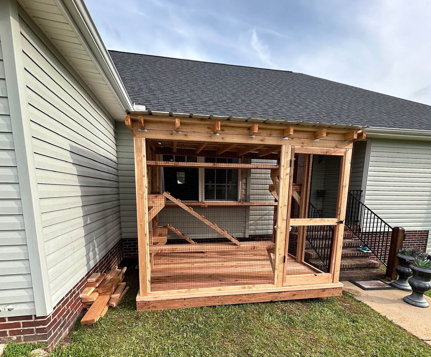 A custom-built wooden catio attached to a light gray house, featuring a slanted roof, mesh wiring for safety, and an elevated ramp inside.