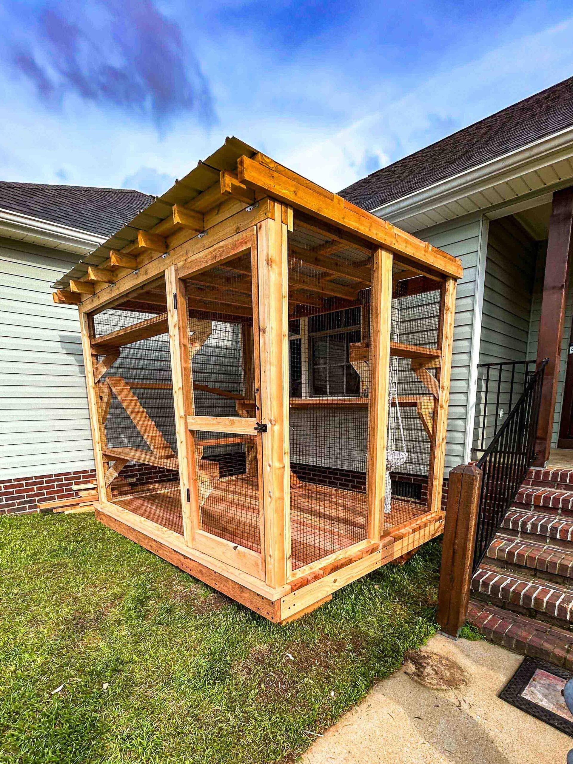 A completed custom catio attached to a home, featuring a slanted wooden roof, secure wire mesh sides, wood flooring, and a visible hanging cat bed inside.