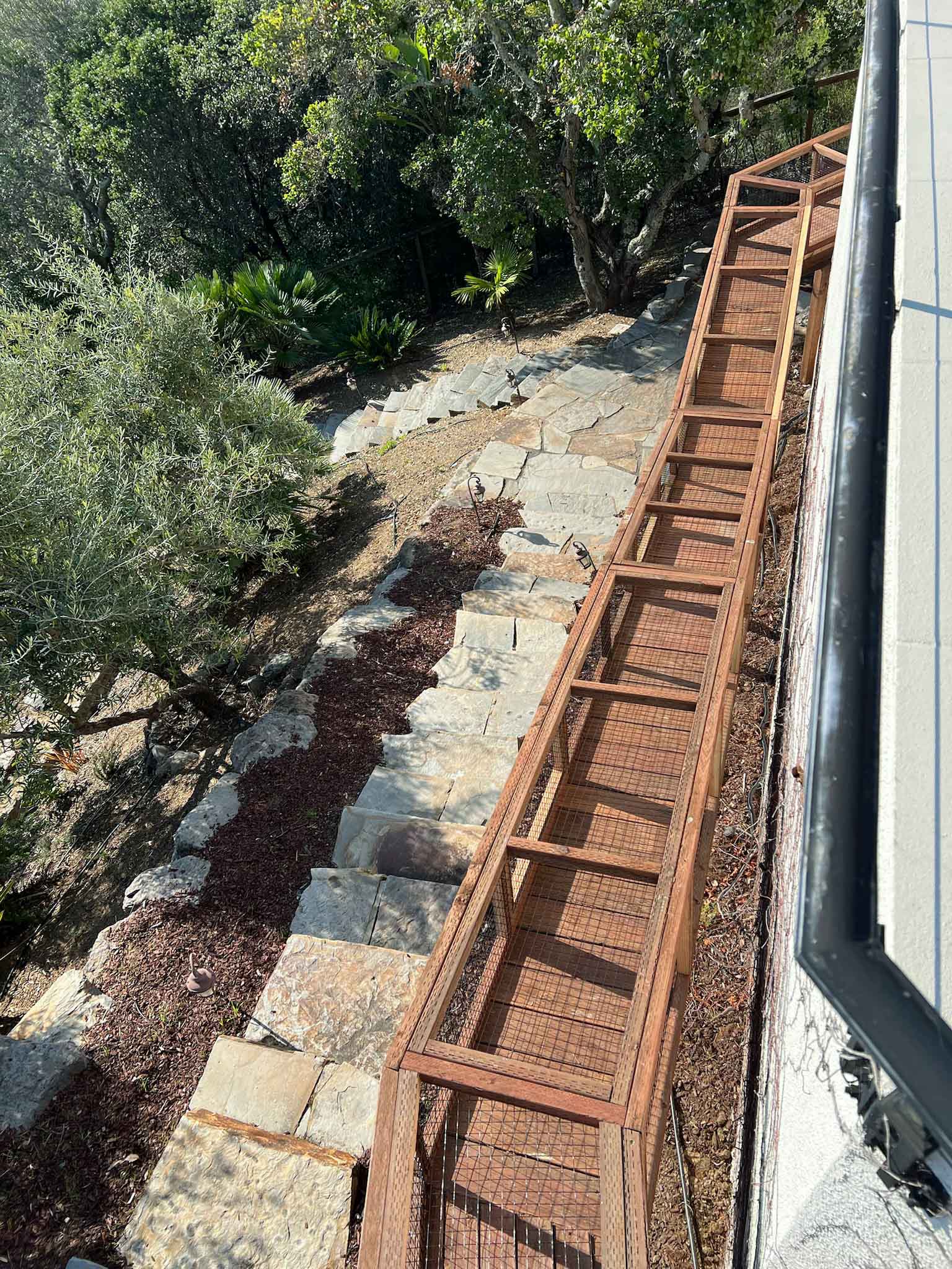 Top-down view of a long, enclosed wooden catio tunnel running alongside a house and descending a hillside landscape.