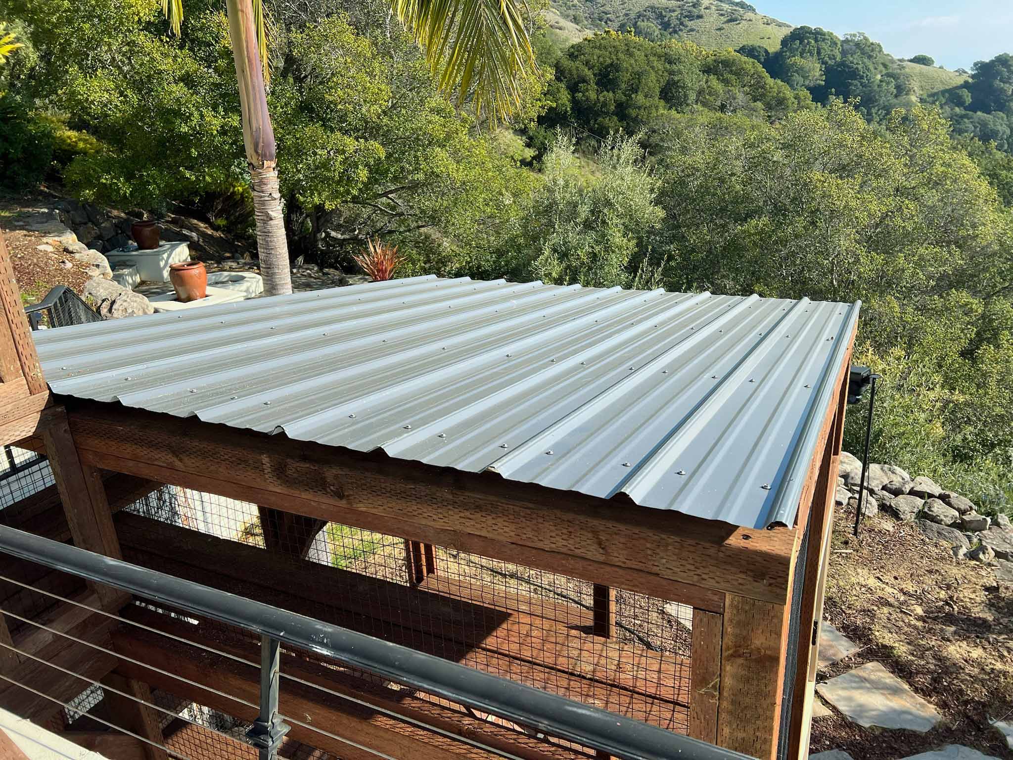 Overhead view of a custom wooden catio featuring a corrugated metal roof, situated on a hillside with a lush forested backdrop.