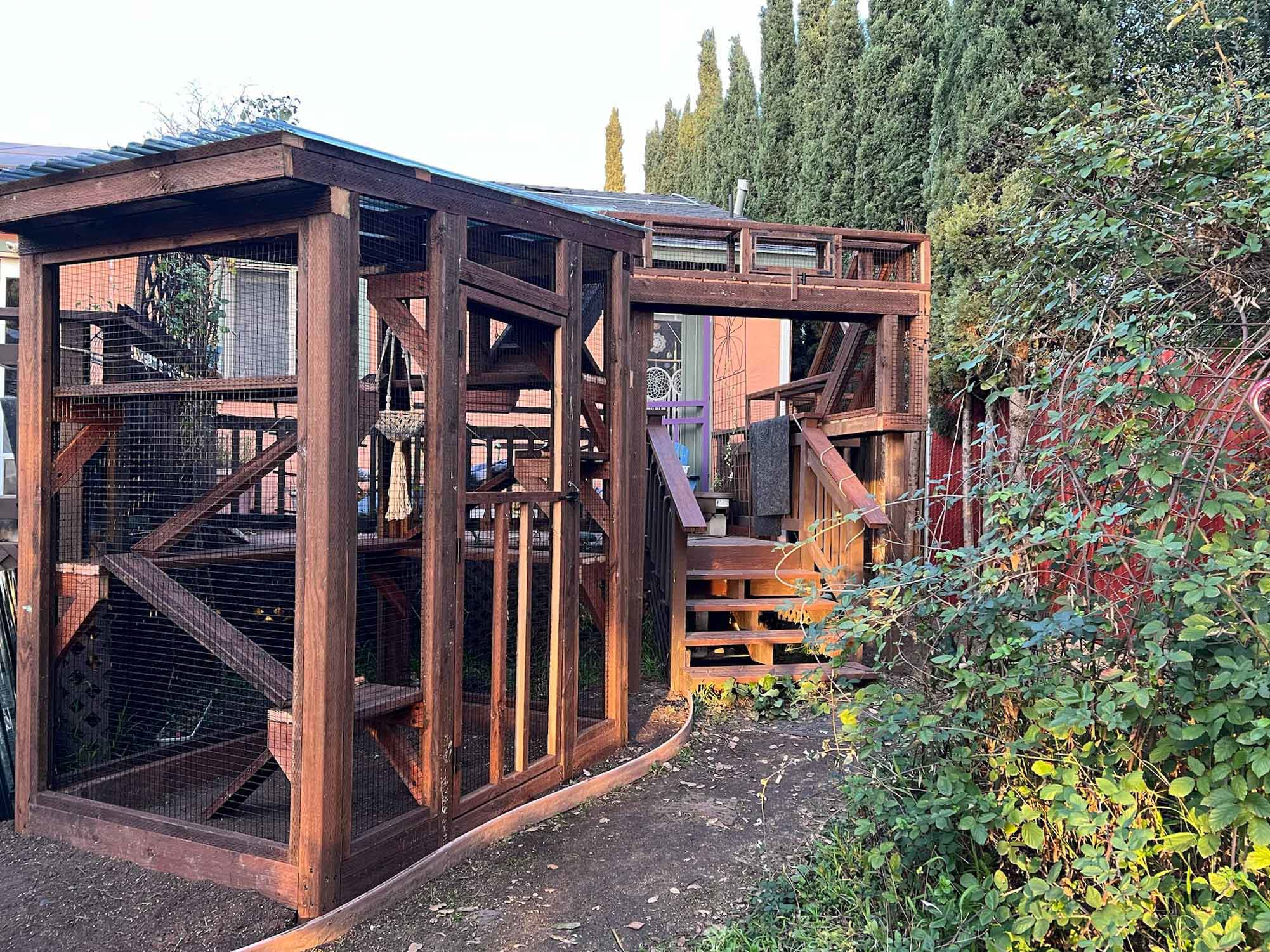 Wide-angle view of a dark wood catio with a roofed enclosure, interior platforms, hanging hammock, and a bridge tunnel leading from a raised deck to the house.