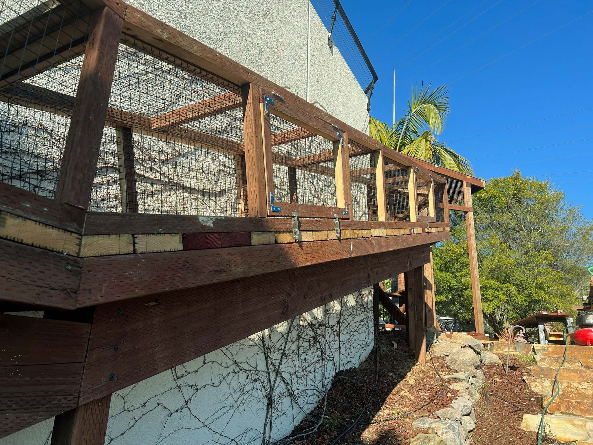Angled side view of an elevated catio tunnel with mesh siding and wooden framing, running along the house above a stone-lined path.