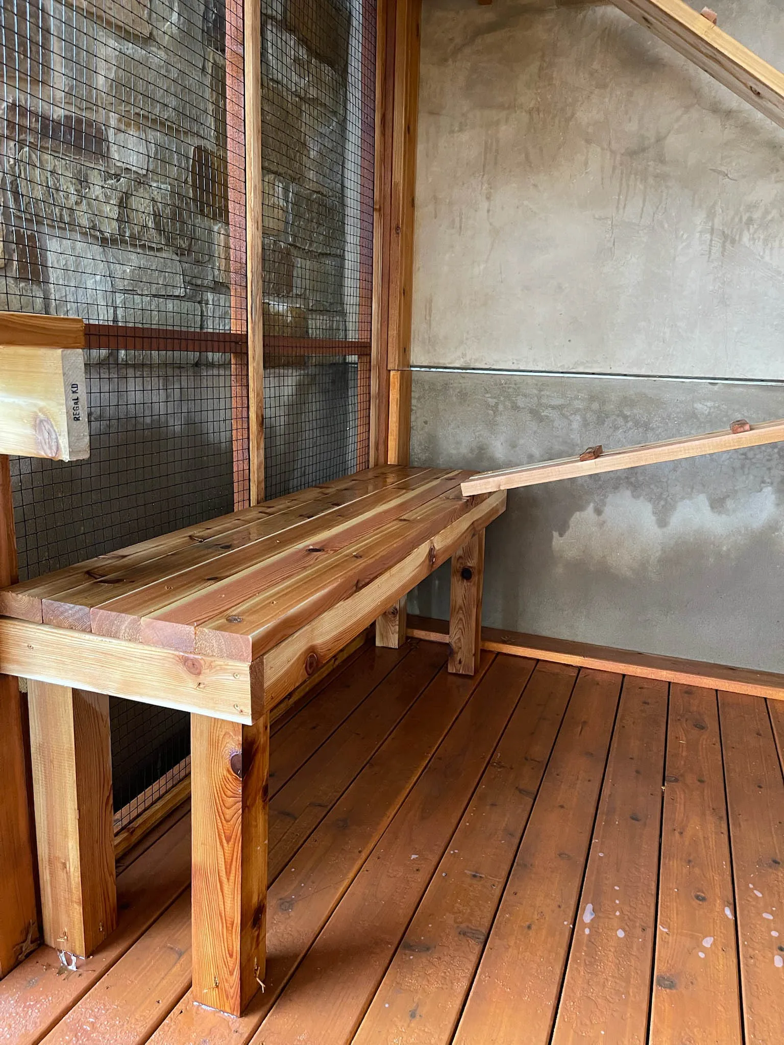 Close-up of a wooden bench inside a catio with mesh walls and a climbing ramp leading upward.