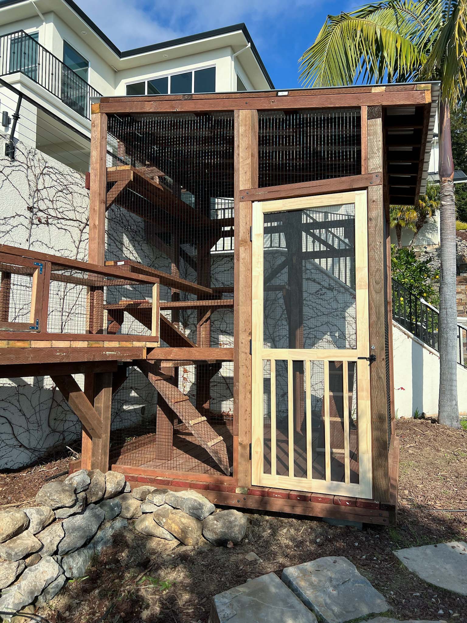 Close-up of a wooden catio entrance with a mesh screen door, elevated platforms inside, and a ramp leading into the structure from the left.