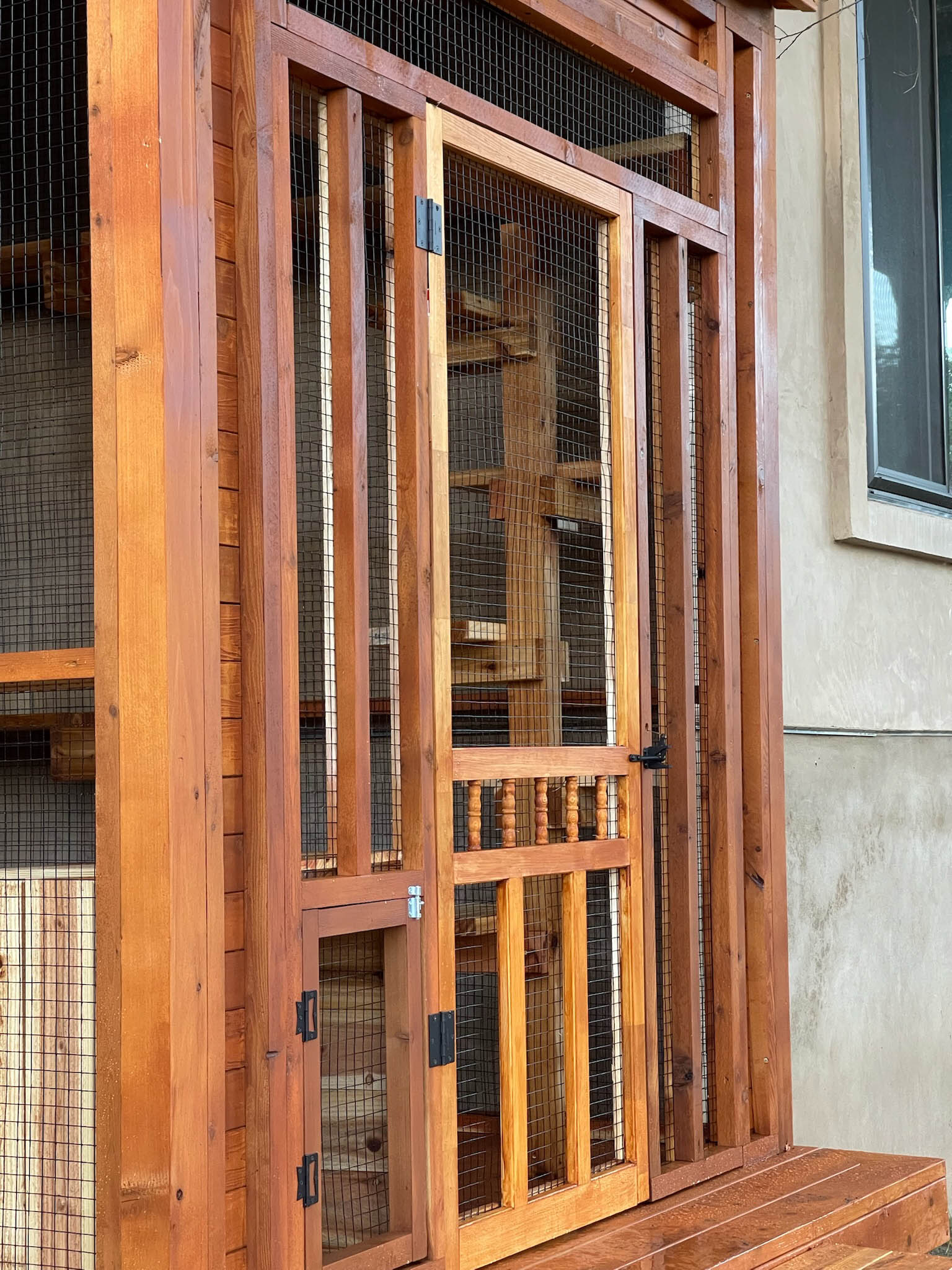 Close-up of a wooden catio entry with a mesh door, decorative spindle detail, and a small latched lower access door.