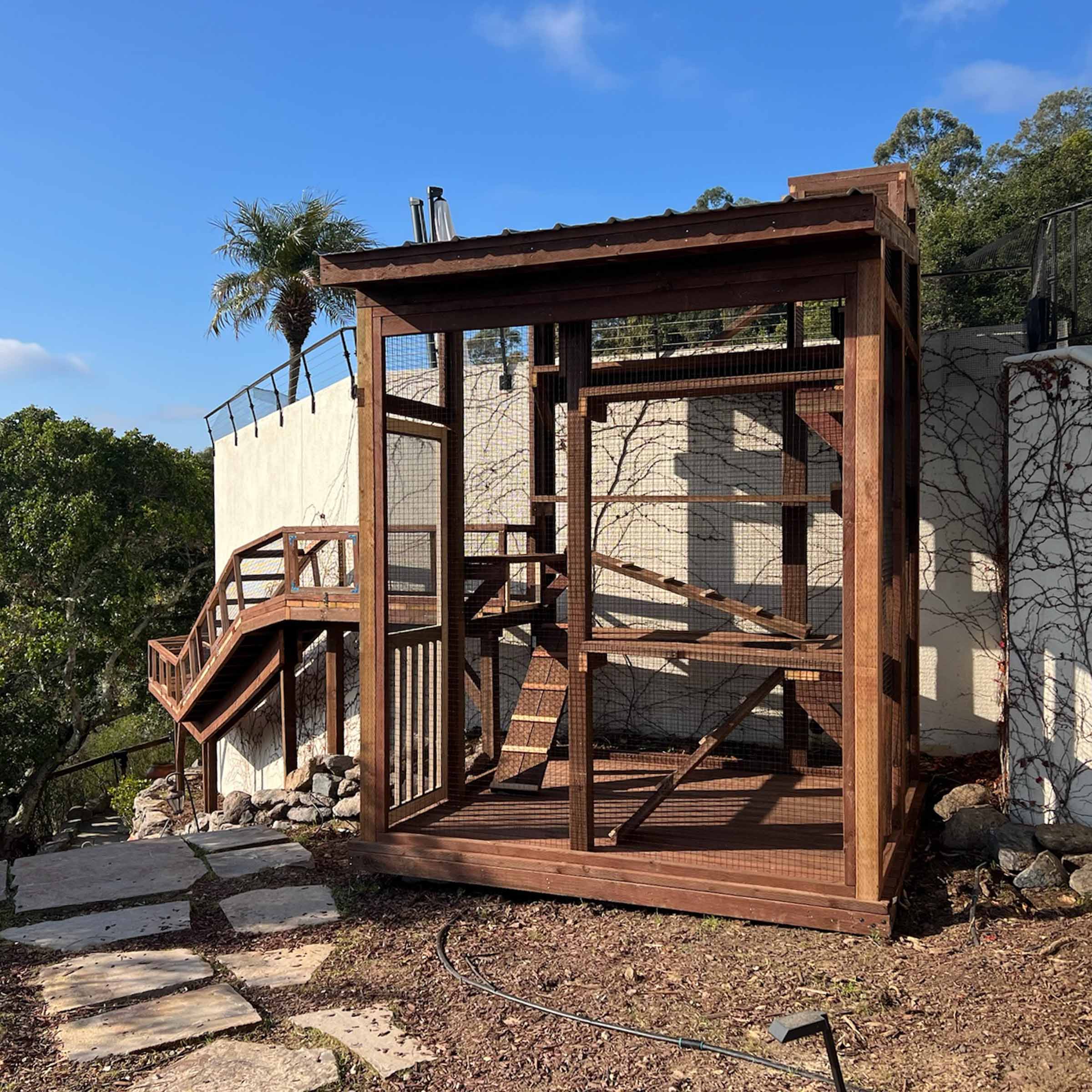 Straight-on view of a large wooden catio enclosure with mesh siding, ramps inside for vertical play, and a stone stepping path leading up to the entrance.