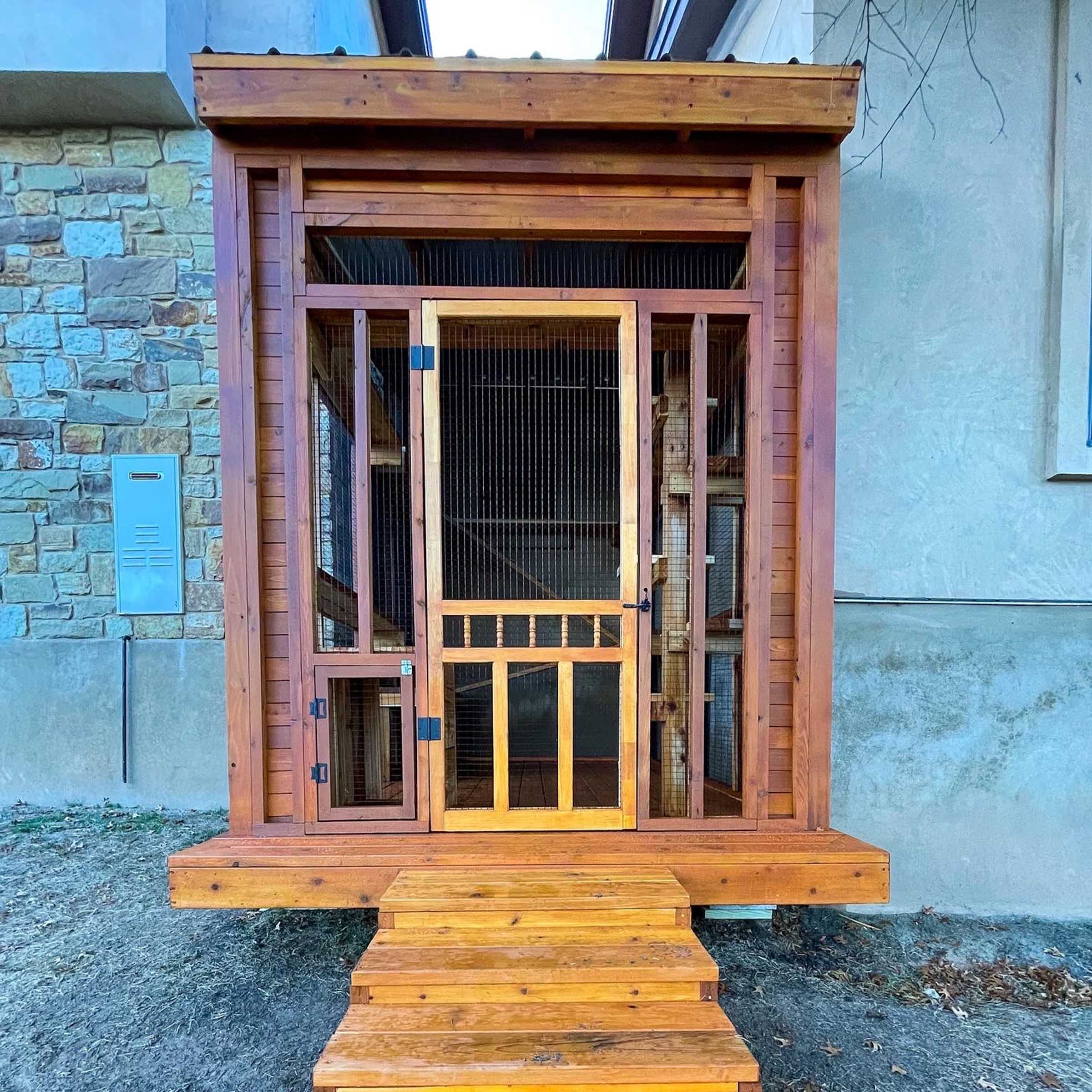 Front-facing view of a raised wooden catio with mesh panels, a main entry door, a secondary lower access panel, and steps leading up.