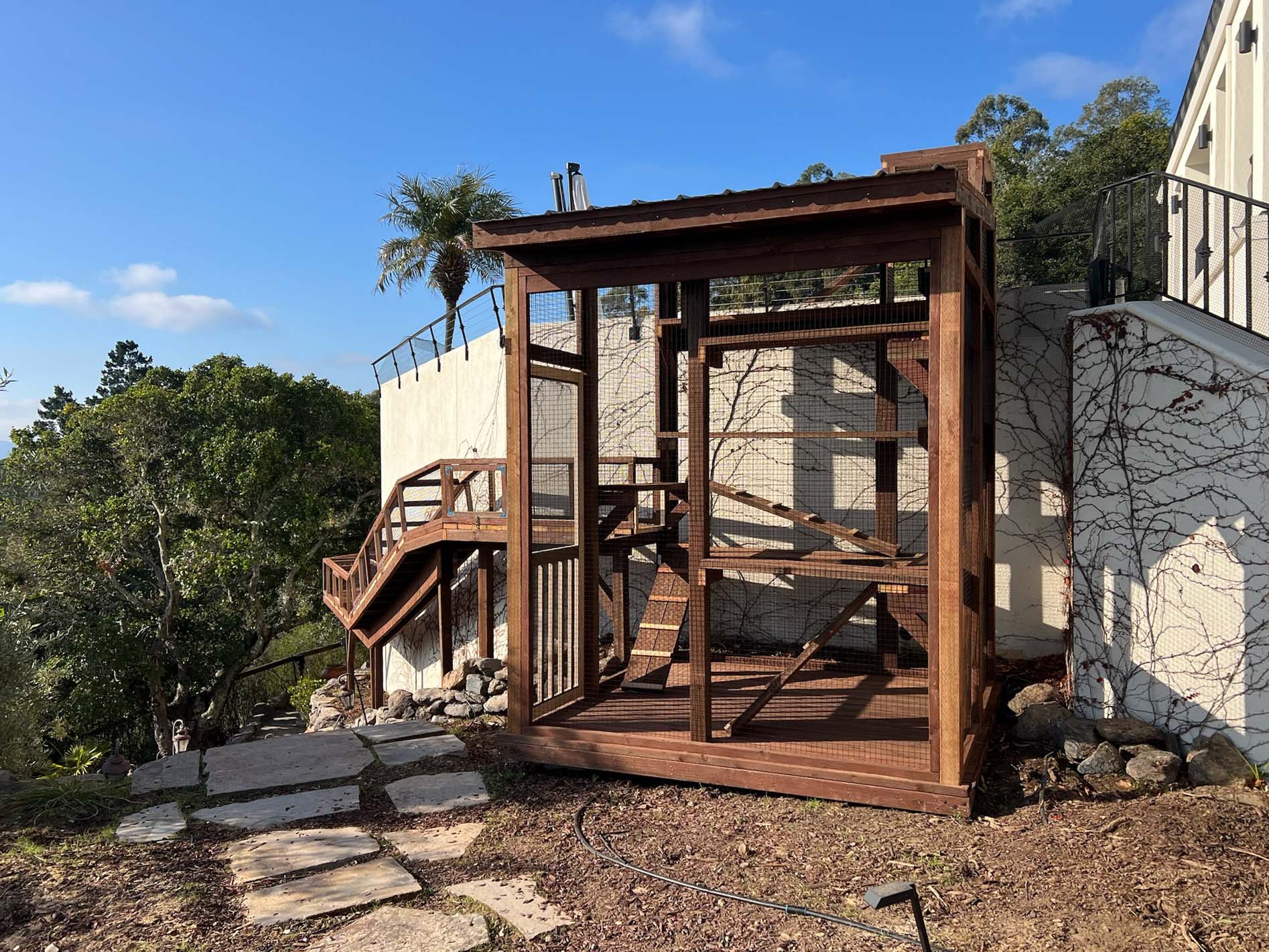 Front view of a multi-tiered catio enclosure attached to a hillside home, featuring internal ramps, mesh walls, and a scenic stone path leading to the structure.