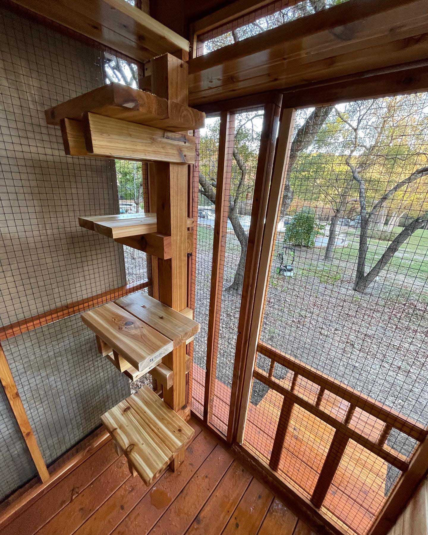 Close-up of wooden cat steps ascending vertically inside a mesh-enclosed catio, with views of trees and grass through the screened panels.