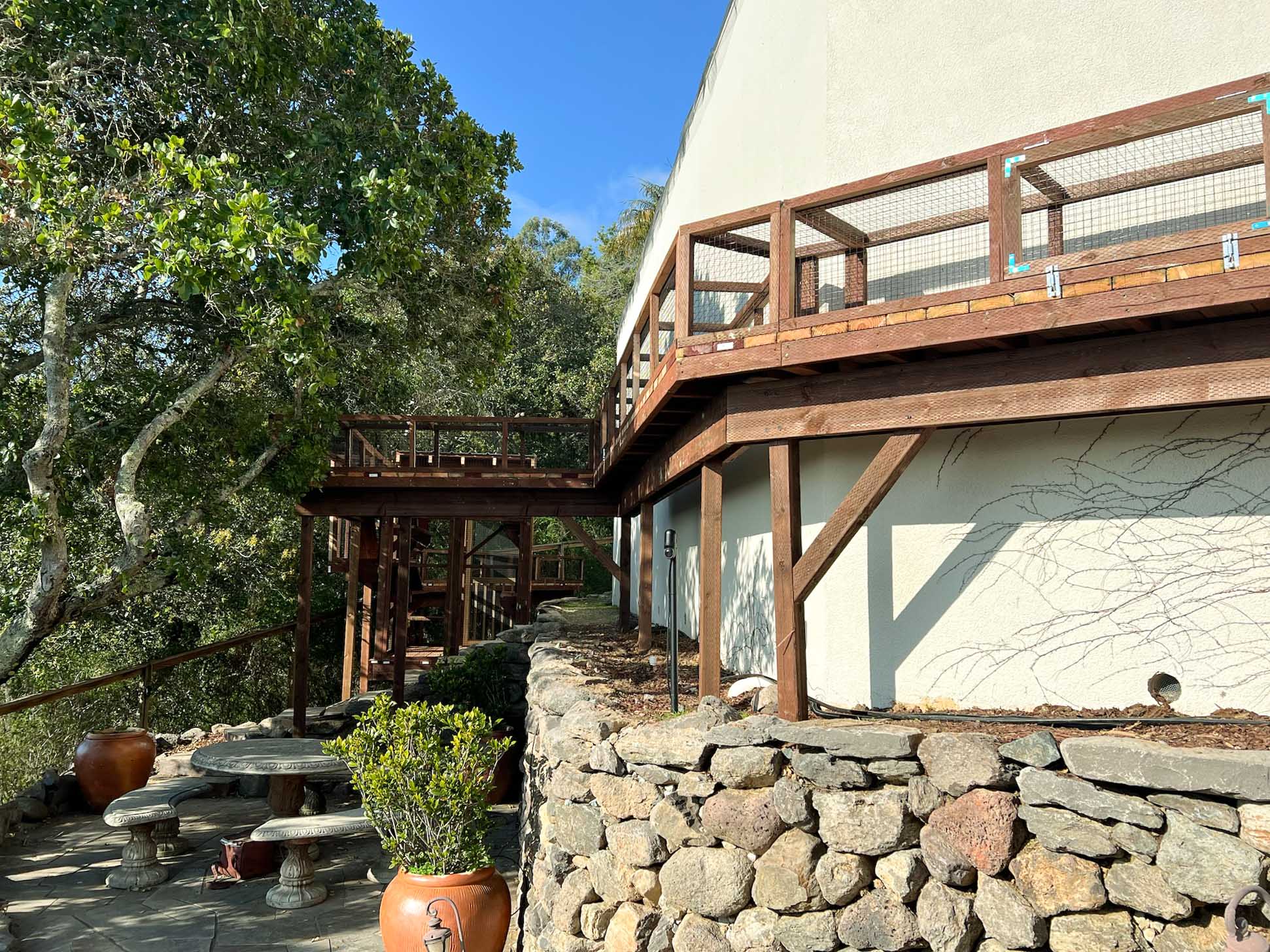 Elevated wooden catio wraps around the side of a house, above a landscaped stone patio area with garden planters and a stone table set under a tree canopy.