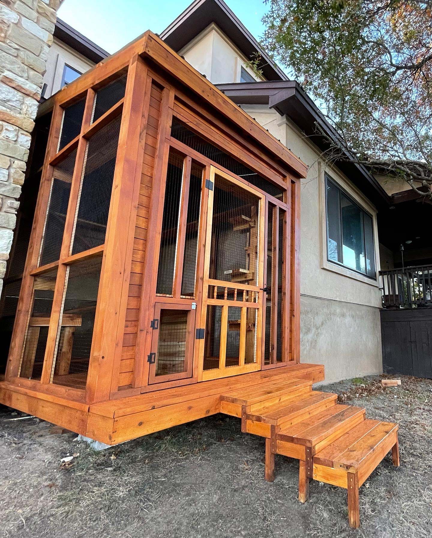 Exterior view of a raised wooden catio with mesh walls, a slanted roof, and a staircase leading to a front entry door.