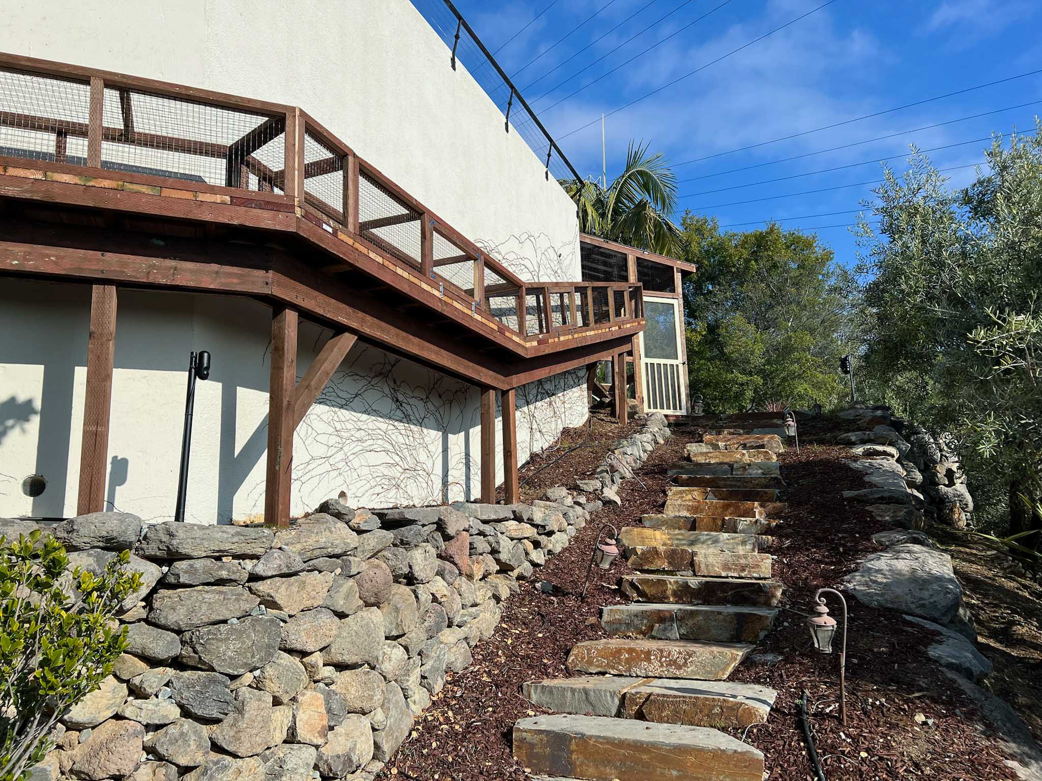 Custom catio with an elevated wooden walkway, enclosed with mesh, stretching alongside a house and connected to a garden path made of stone slab steps.