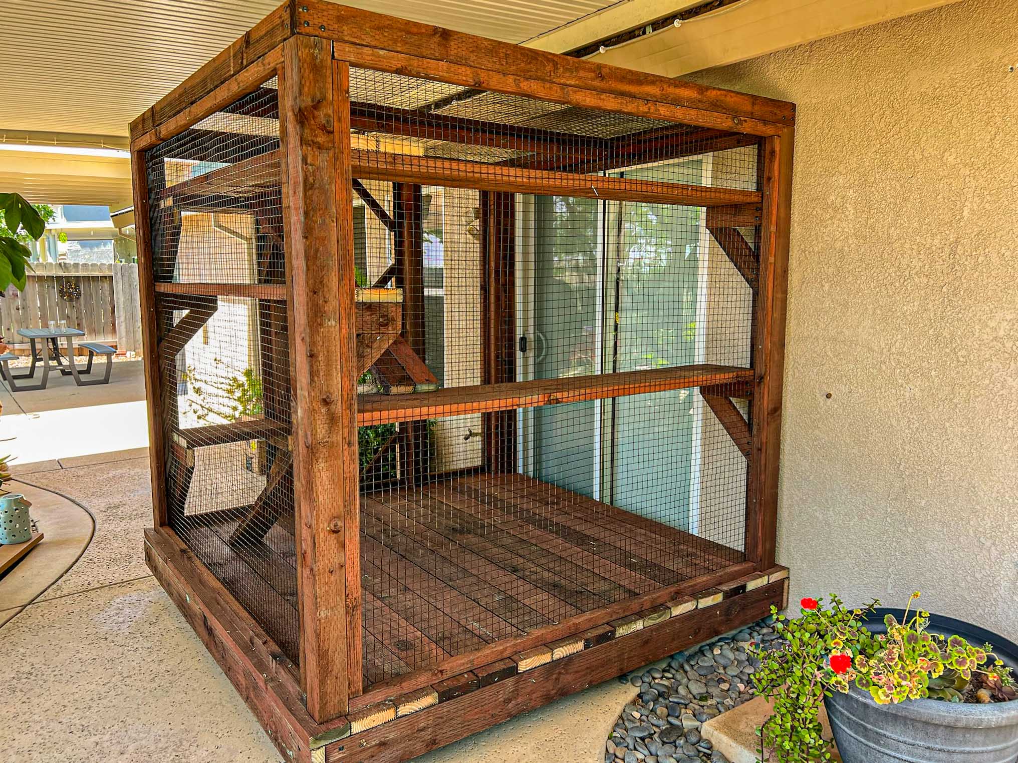 Spacious custom catio made of stained wood and wire mesh, attached to a house with sliding glass doors.