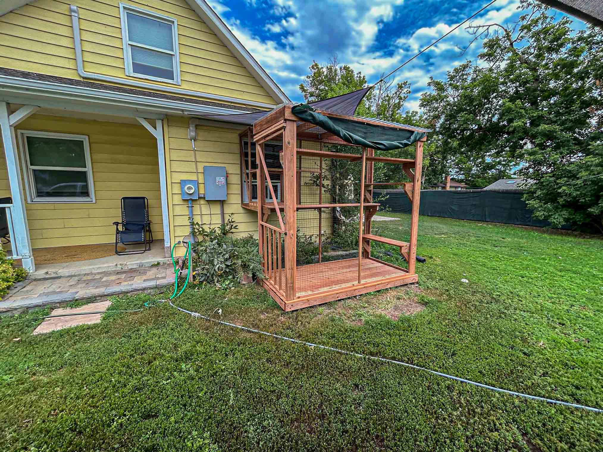 Custom catio with wood framing and green awning, attached to a yellow house beside a small porch and open grassy backyard.