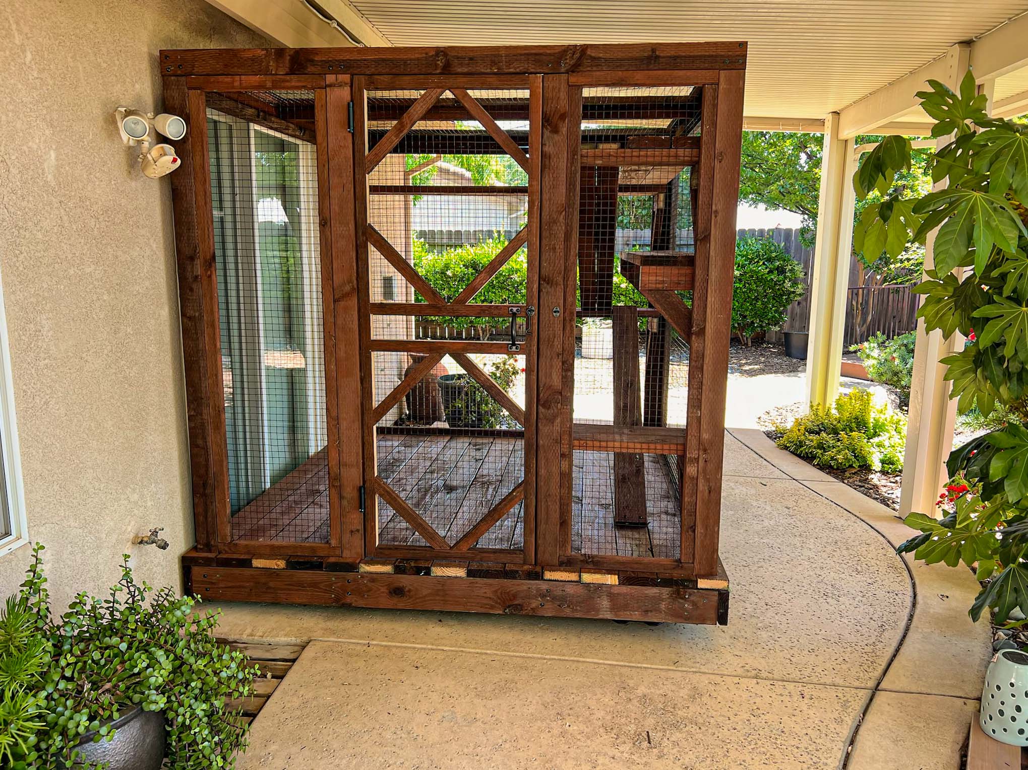 Outdoor wooden catio enclosure attached to a house patio with wire mesh walls and a secure door.