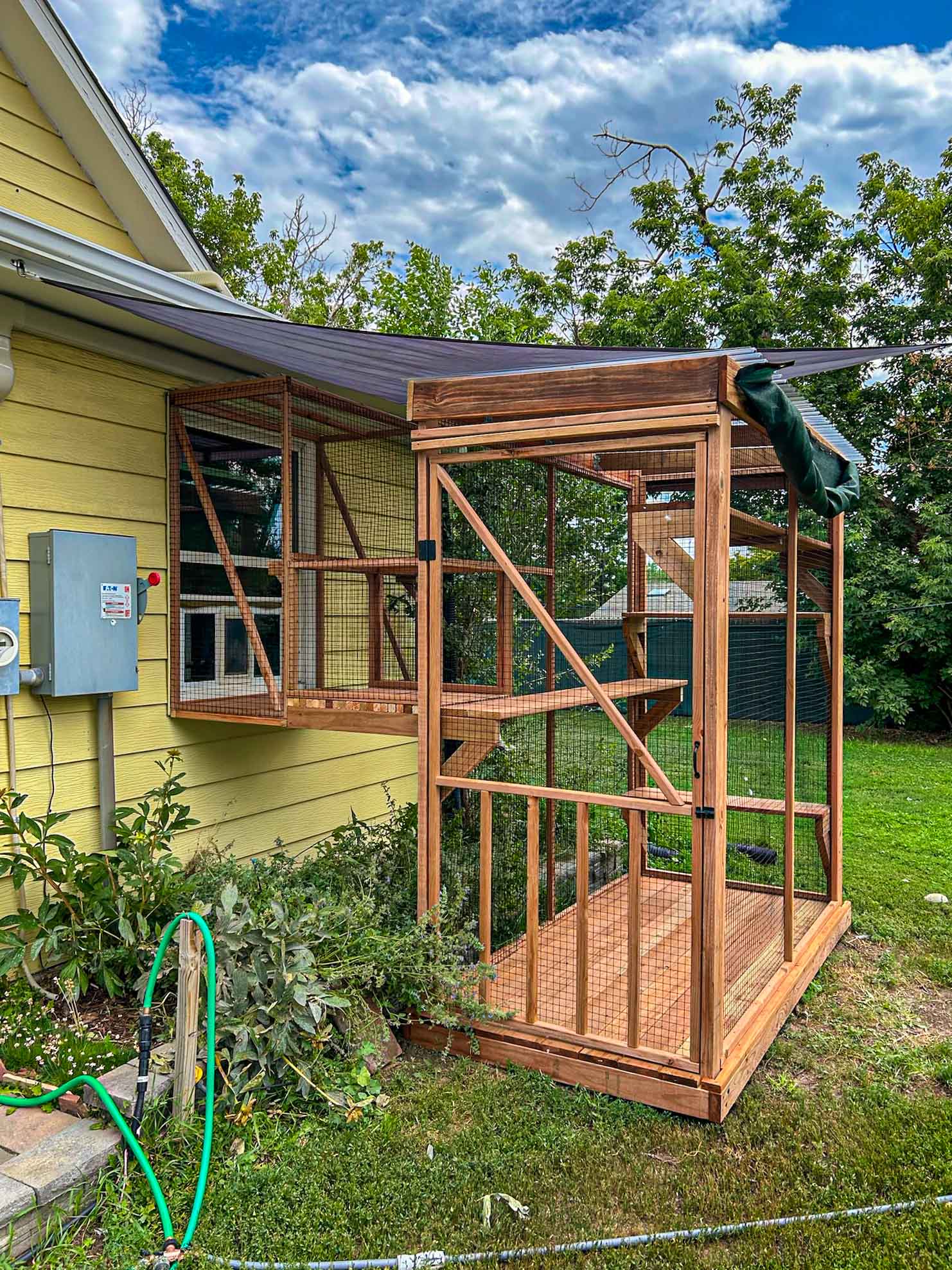 Close-up view of a wooden catio with mesh walls and enclosed window bridge, connected to a yellow house with a dark overhead awning.