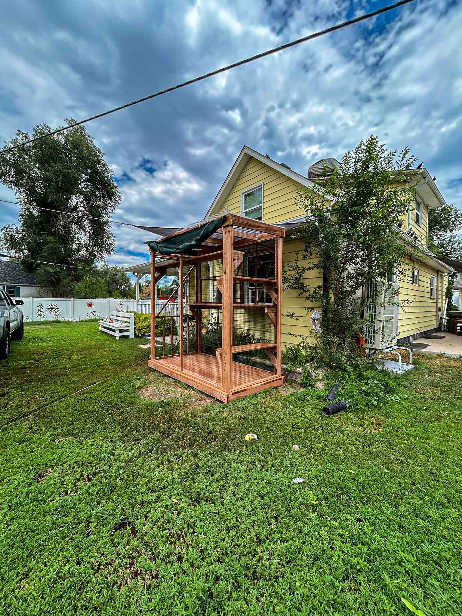 Angled view of a wooden catio with mesh panels, shaded awning, and vertical layout, attached to a yellow two-story home in a grassy backyard.