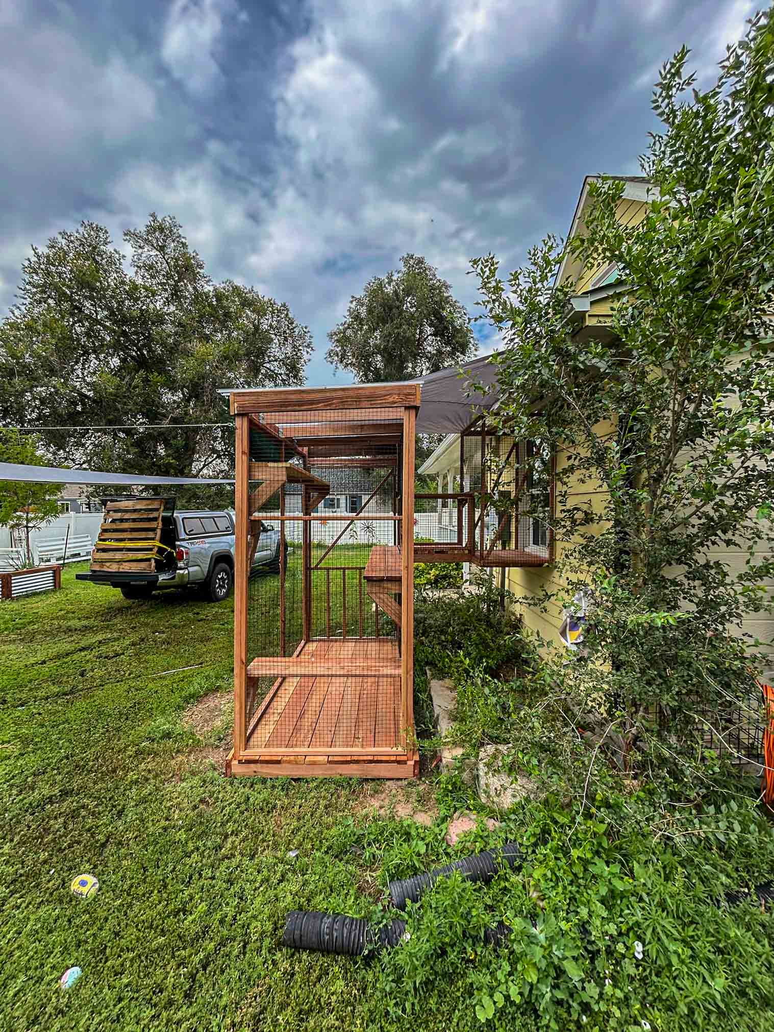 Front-facing view of a wooden catio with mesh walls, raised perch, and ramp, connected to a house window with a shaded awning overhead.