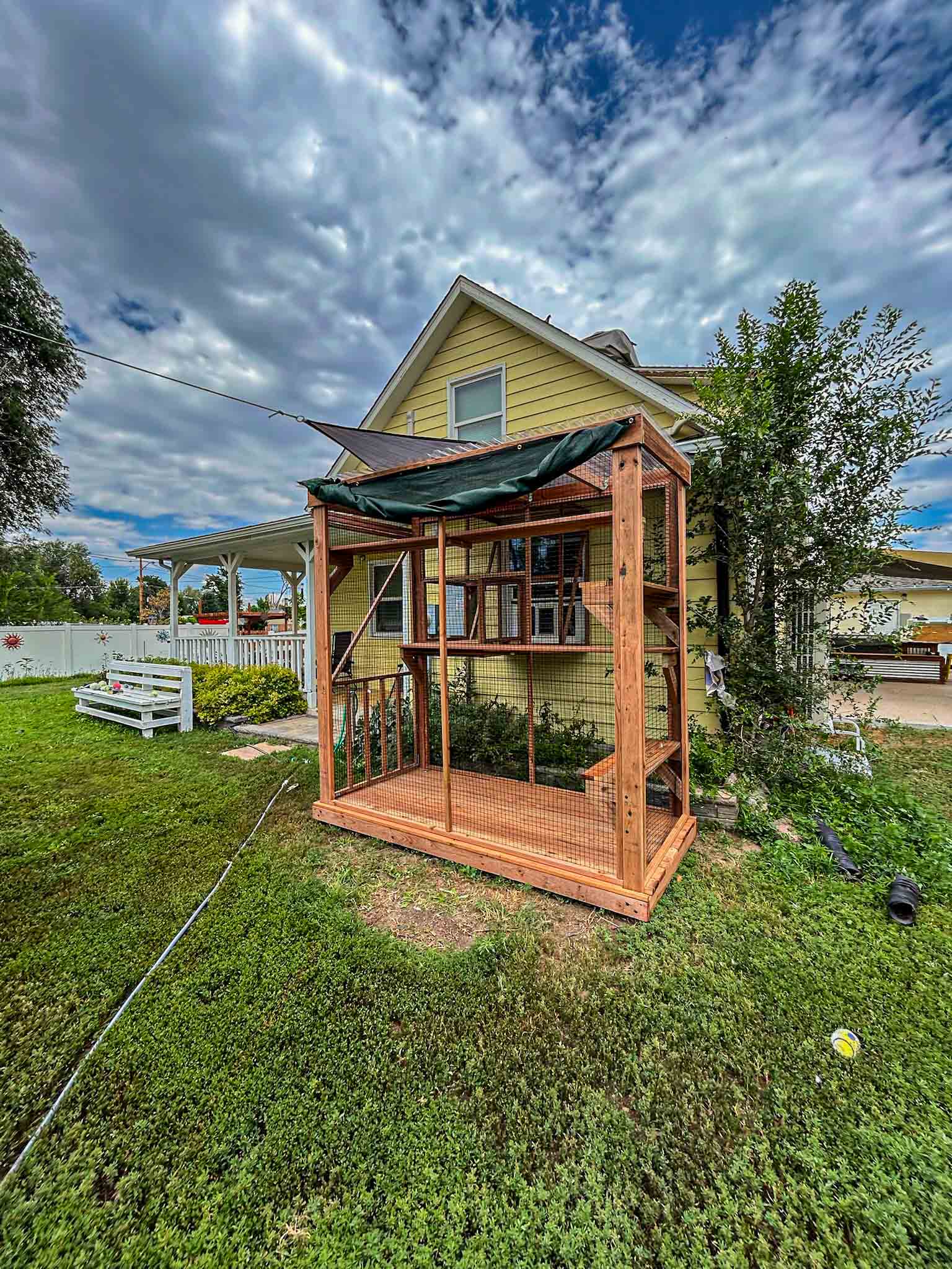 Wide-angle view of a tall wooden catio with green awning, attached to a yellow house and surrounded by lush green lawn and garden.