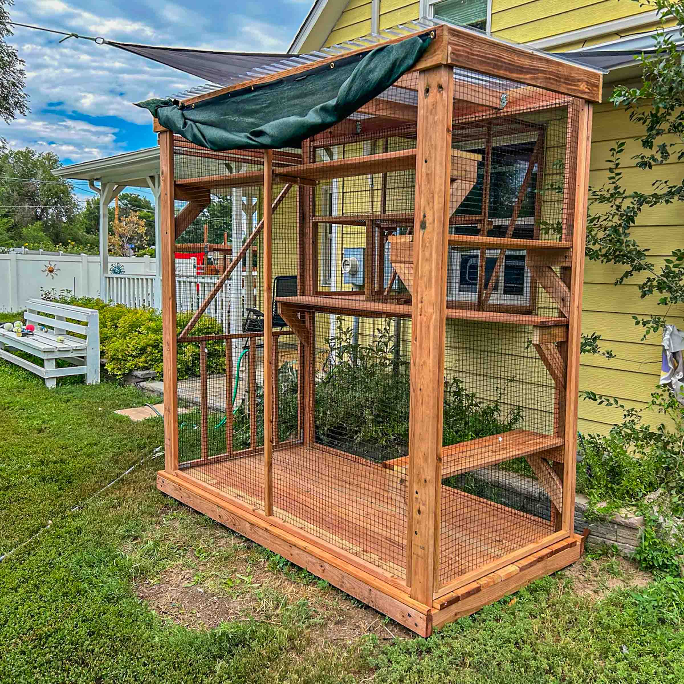 Side view of a vertical wooden catio with multiple platforms and mesh walls, positioned against a yellow house with a green awning overhead.