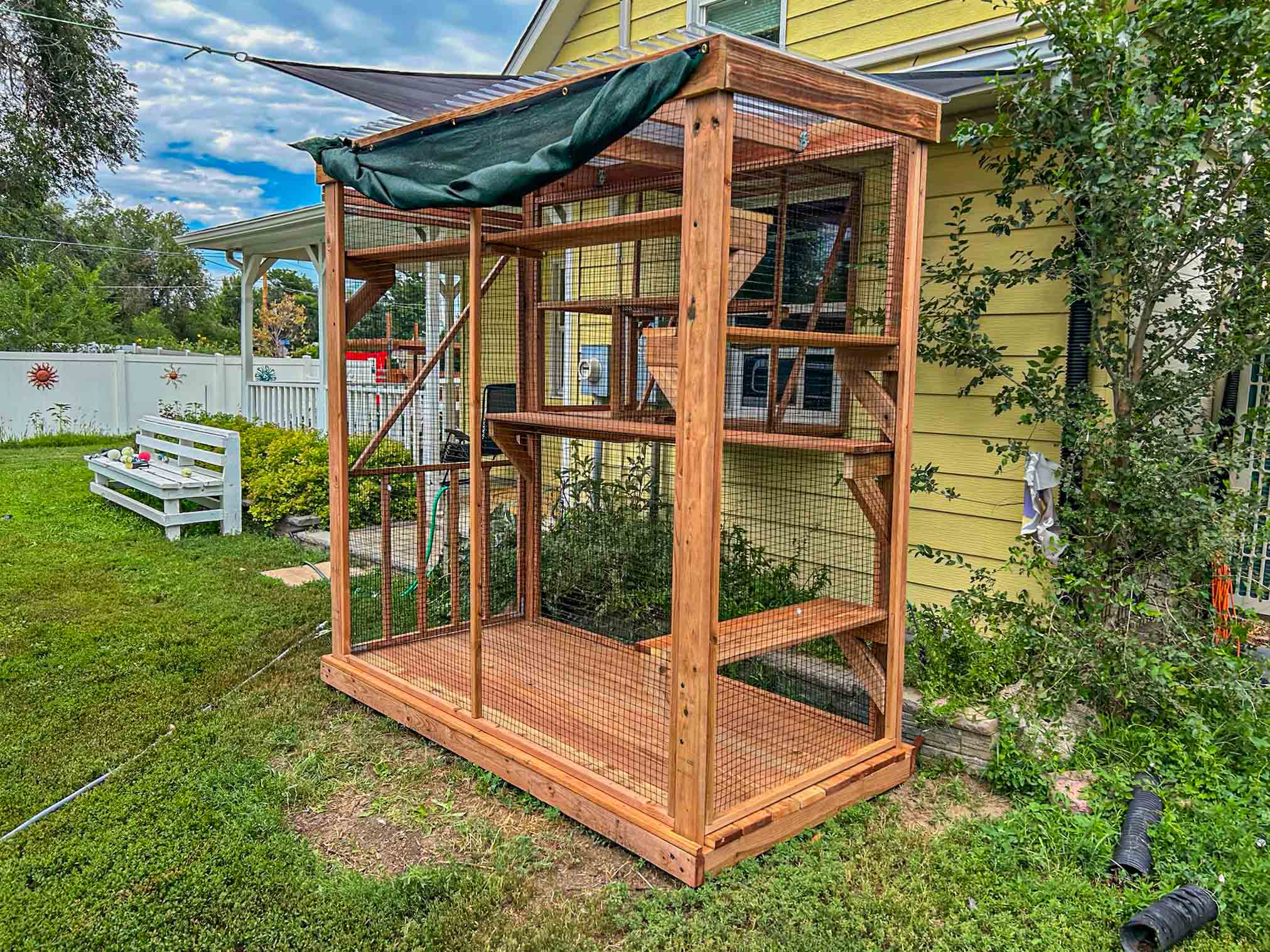 Freestanding vertical wooden catio with mesh sides and multiple perches, attached to a yellow house with surrounding garden and lawn.
