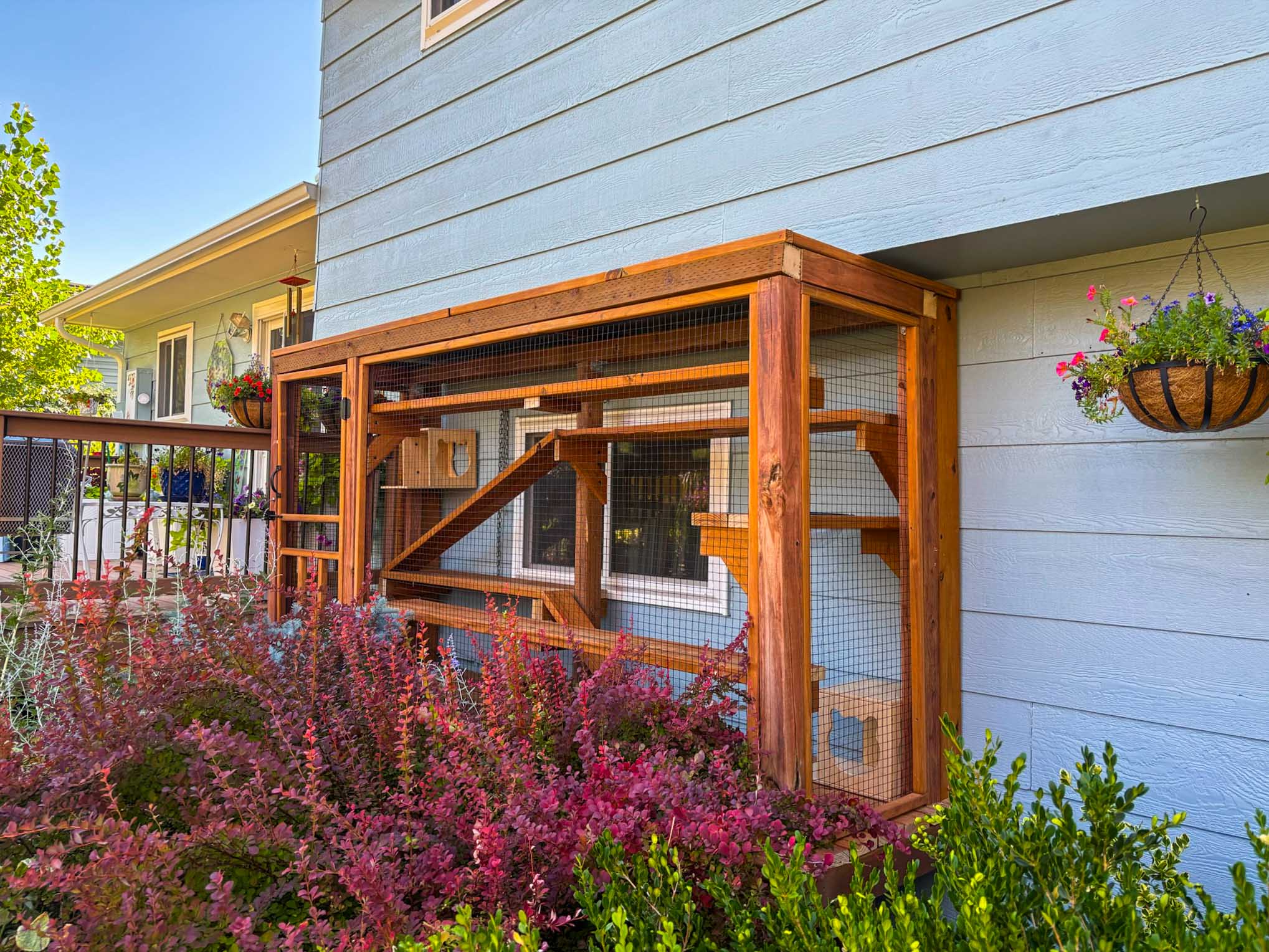 A custom-built wooden catio attached to a house exterior, surrounded by vibrant landscaping and colorful hanging flower baskets.