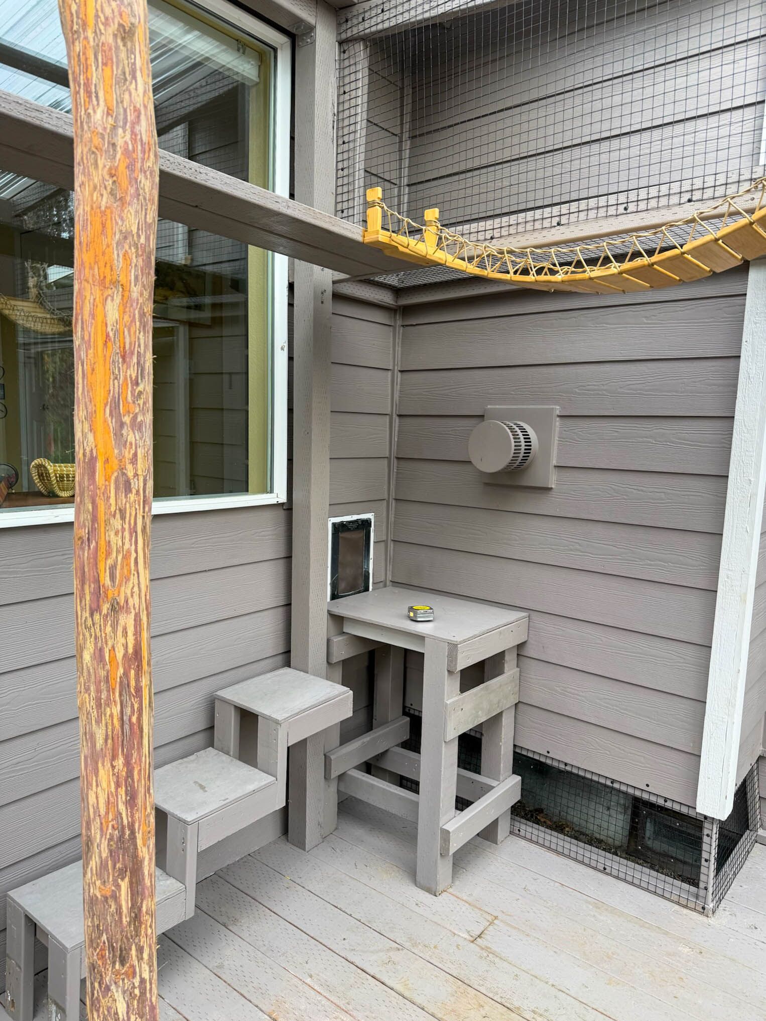 Corner section of a catio with a small cat door, steps, and a yellow bridge leading to an upper platform.