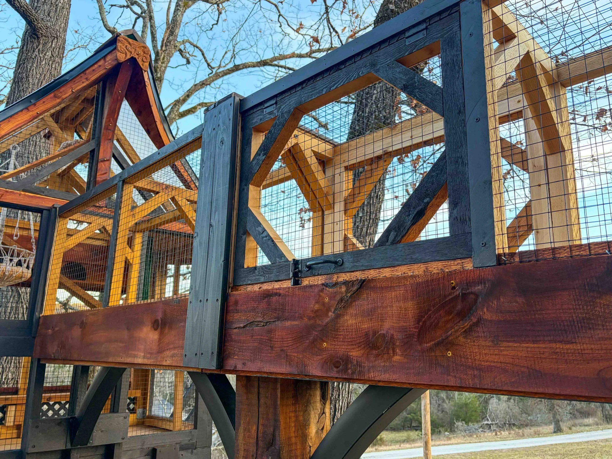 Detailed side view of a custom-built wooden catio enclosure with dark trim, mesh railing panels, and a sloped roof section framed in cedar, captured from a low upward angle.