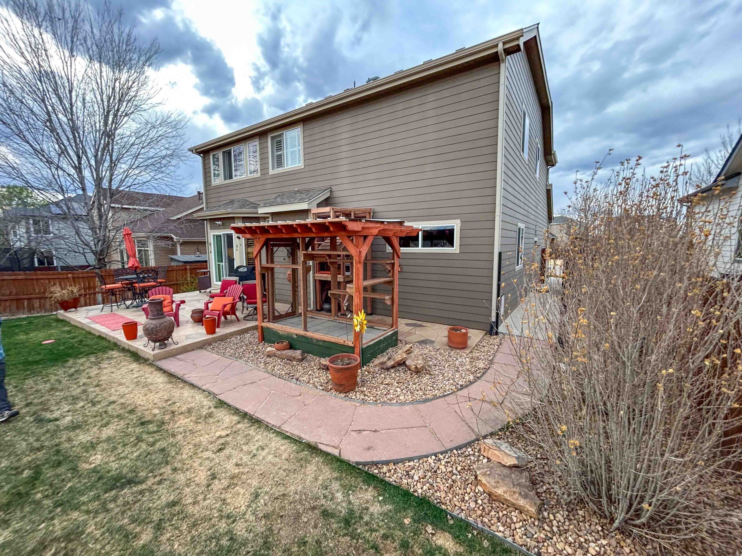 Wide view of backyard with custom-built catio enclosure attached to house, featuring pergola roof, climbing platforms, and landscaped surroundings with stone walkway and patio furniture.