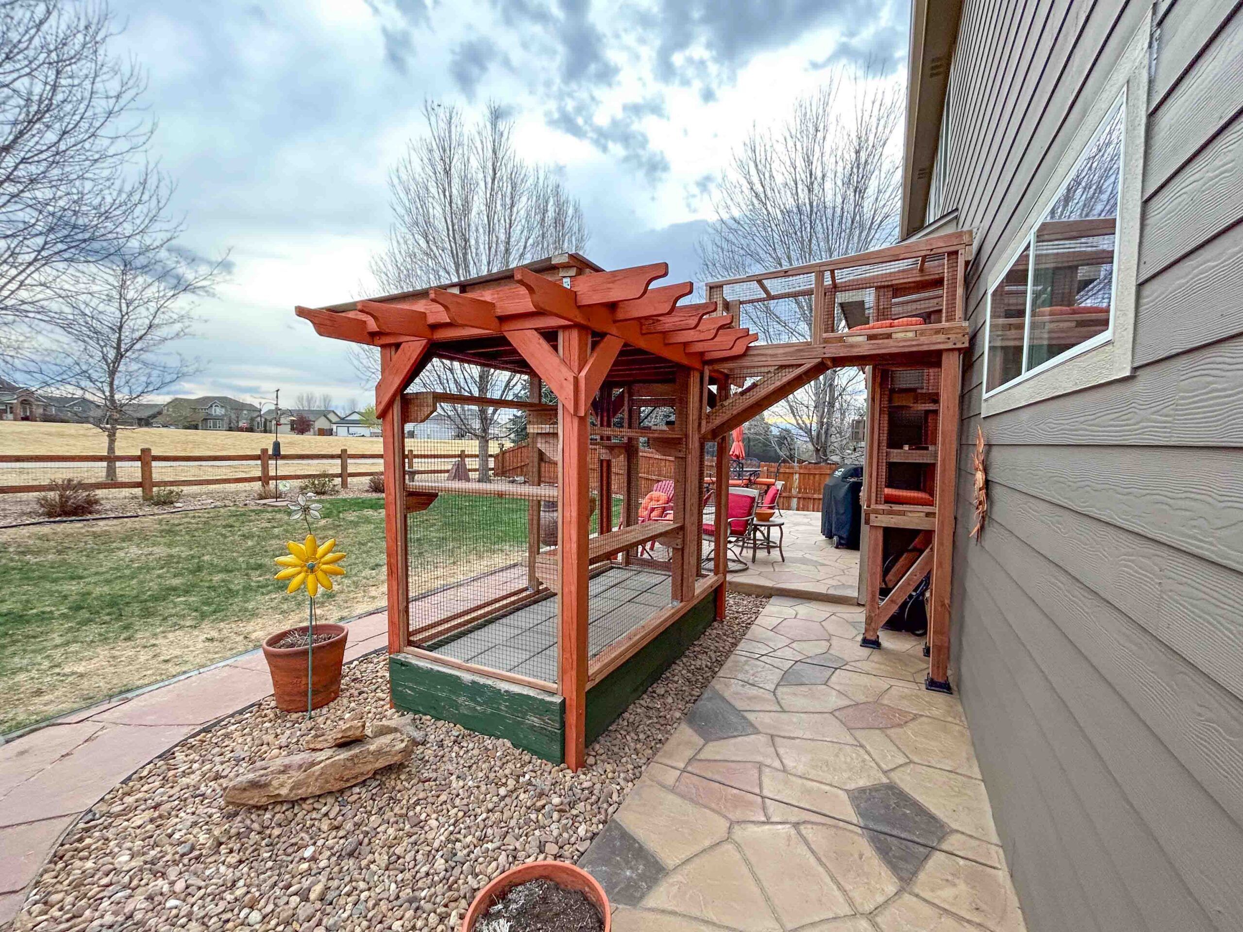 Side view of custom cedar catio with pergola roof and elevated cat tunnel leading to a multi-level climbing tower, positioned along a stone patio beside the home.