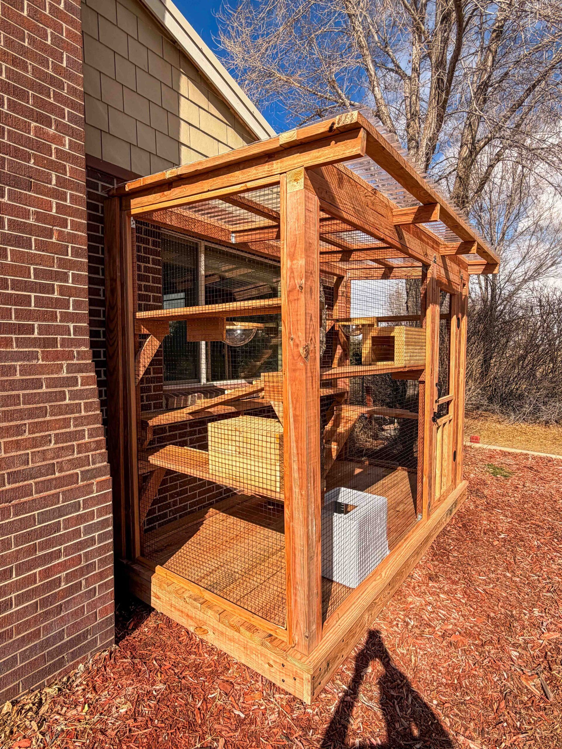 Side view of a custom-built wooden catio attached to a brick home showing cedar posts, wire mesh walls, a slanted clear roof panel, and multiple interior cat shelves and climbing platforms.