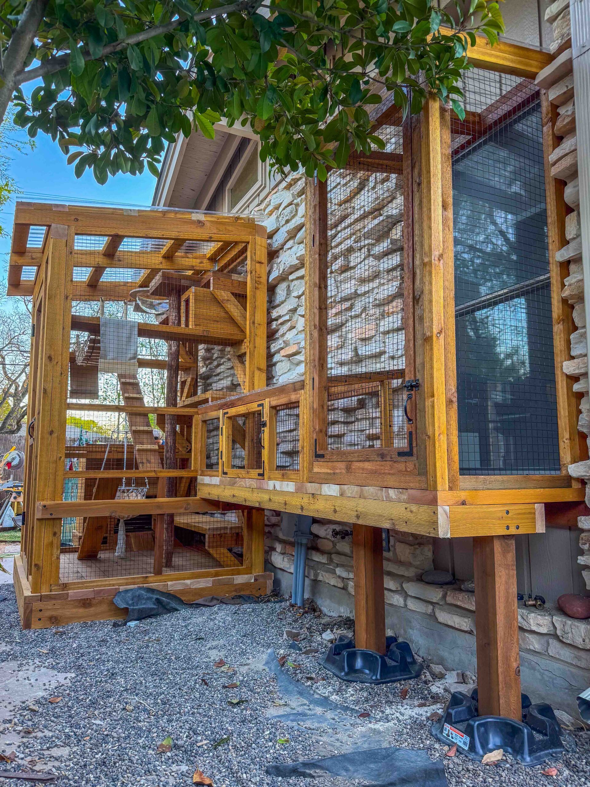 Exterior view of a large custom catio attached to a home with elevated window access, wooden framing, wire mesh panels, and a connected enclosure extension.
