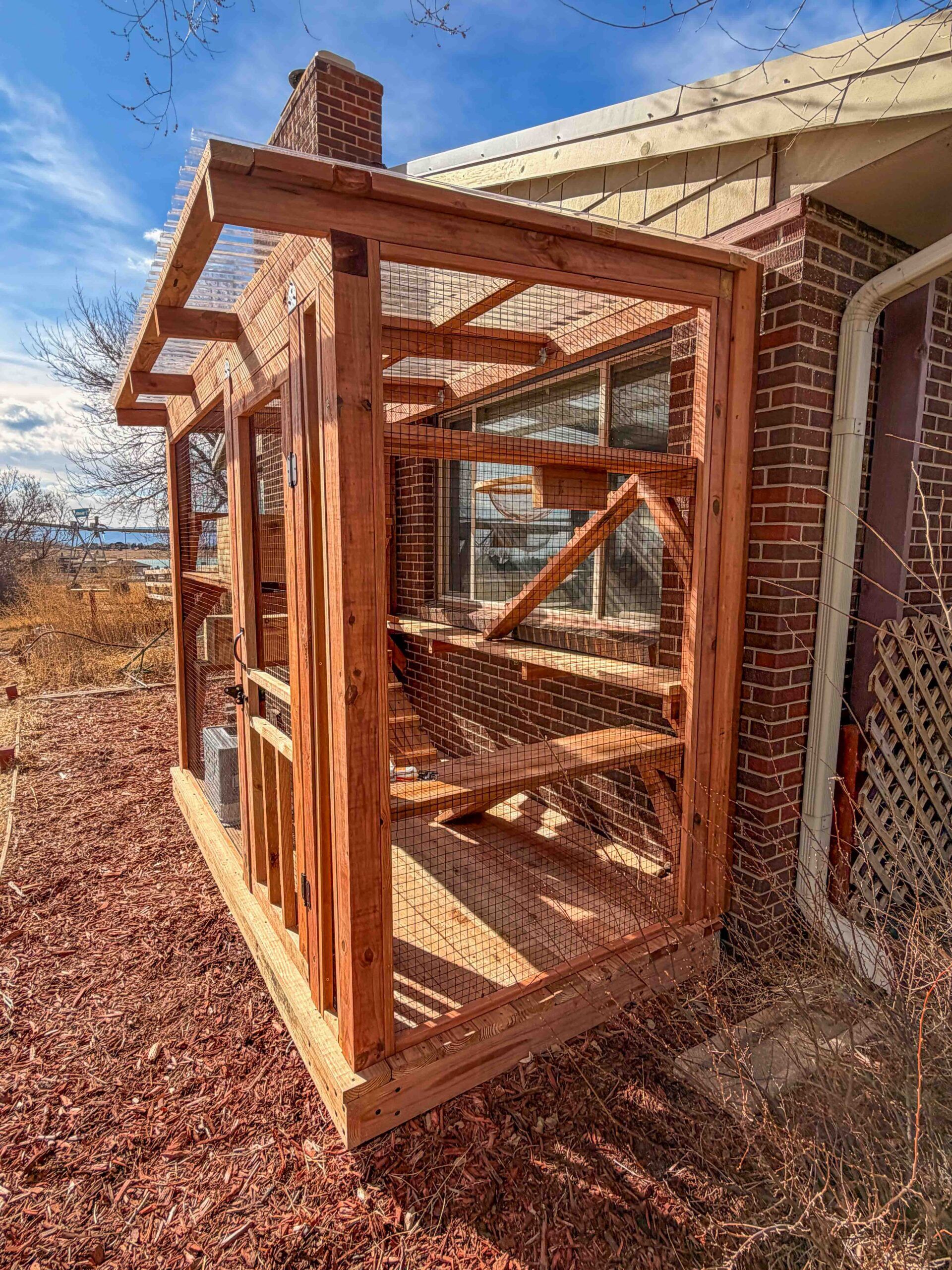 Large custom wooden catio attached to a brick house featuring cedar framing, wire mesh enclosure, multiple interior cat shelves and ramps, and a clear corrugated roof panel for weather protection and sunlight.