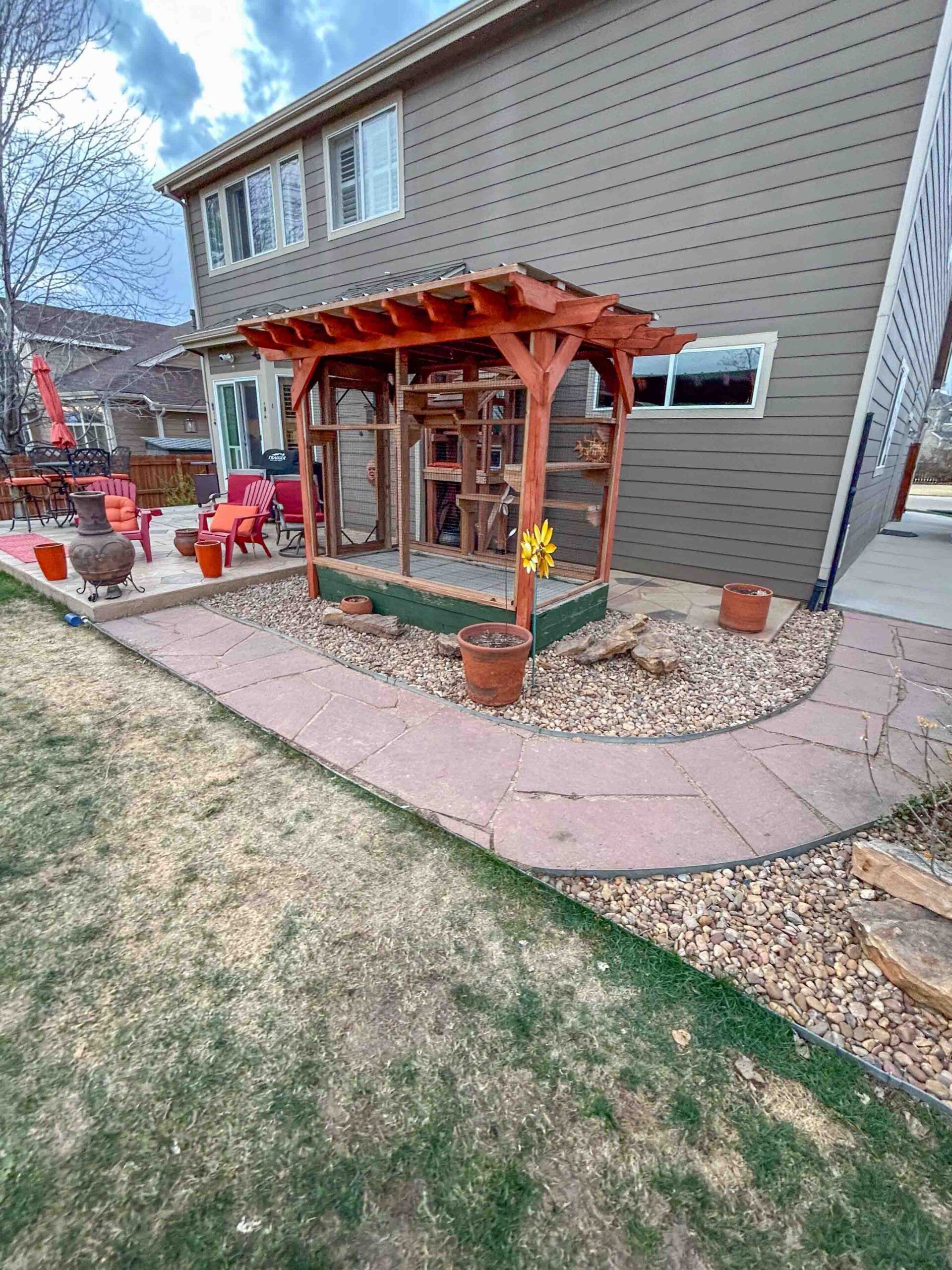 Large custom backyard catio with cedar wood framing and pergola-style roof attached to a two-story home, featuring multiple interior shelves, climbing structures, and a secure enclosed design surrounded by a landscaped patio with stone walkway and outdoor seating.