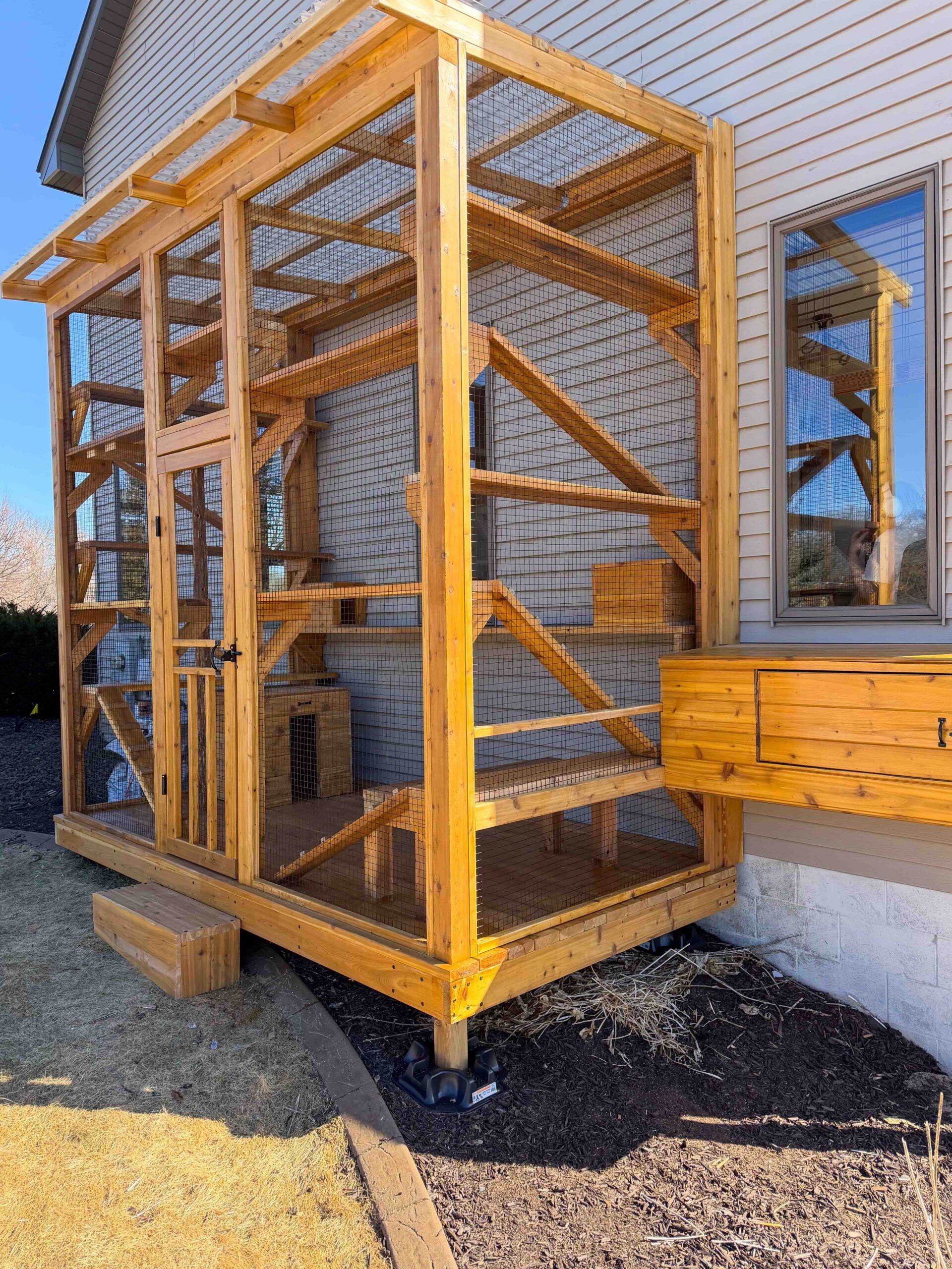 Large custom-built wooden catio attached to the side of a house featuring multiple climbing shelves, ramps, and platforms enclosed with secure wire mesh and topped with a partially covered roof for weather protection.