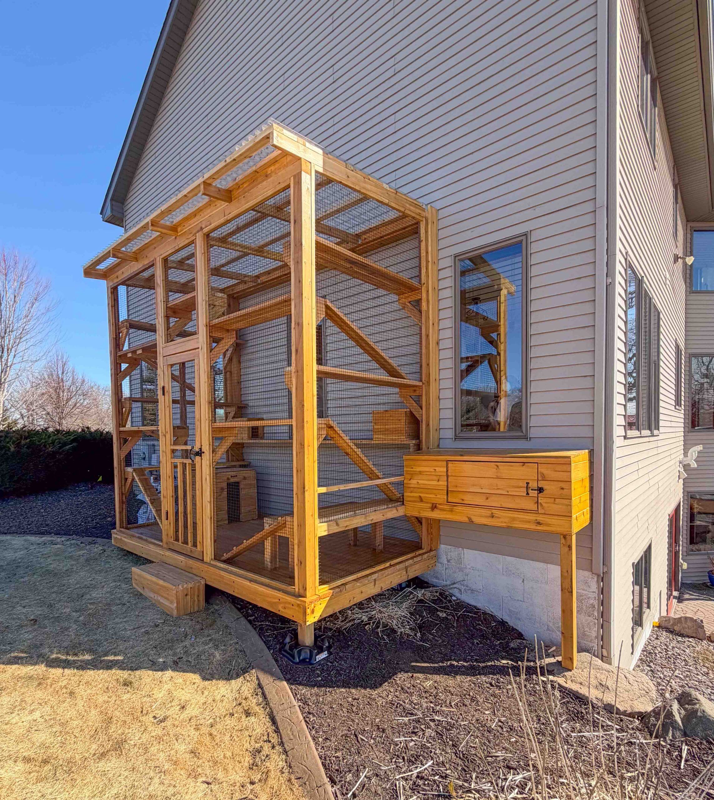 Wide view of a custom-built cedar catio attached to a house with an elevated tunnel connection, wire mesh enclosure, and multiple interior climbing shelves and platforms.