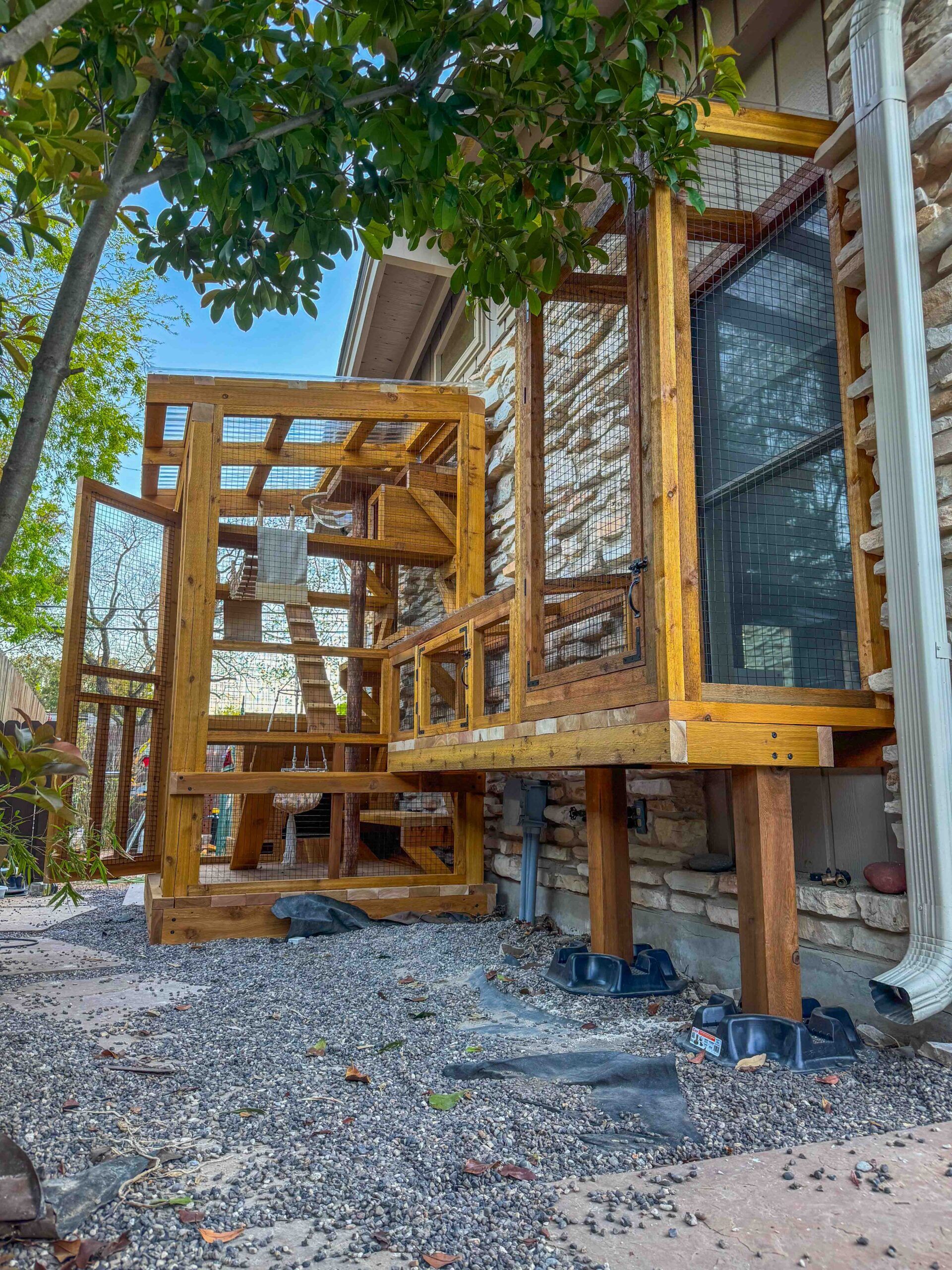 Wide exterior shot of a custom catio attached to a stone house, elevated on posts with a connected enclosure and shaded by a nearby tree.
