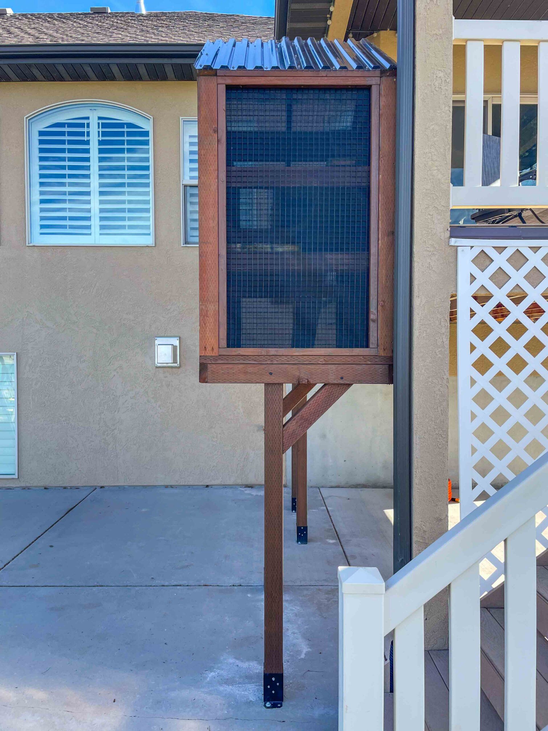 Elevated wooden catio enclosure attached to a beige house exterior, featuring black wire mesh panels, a sloped metal roof, and supported by tall wooden posts over a concrete patio.