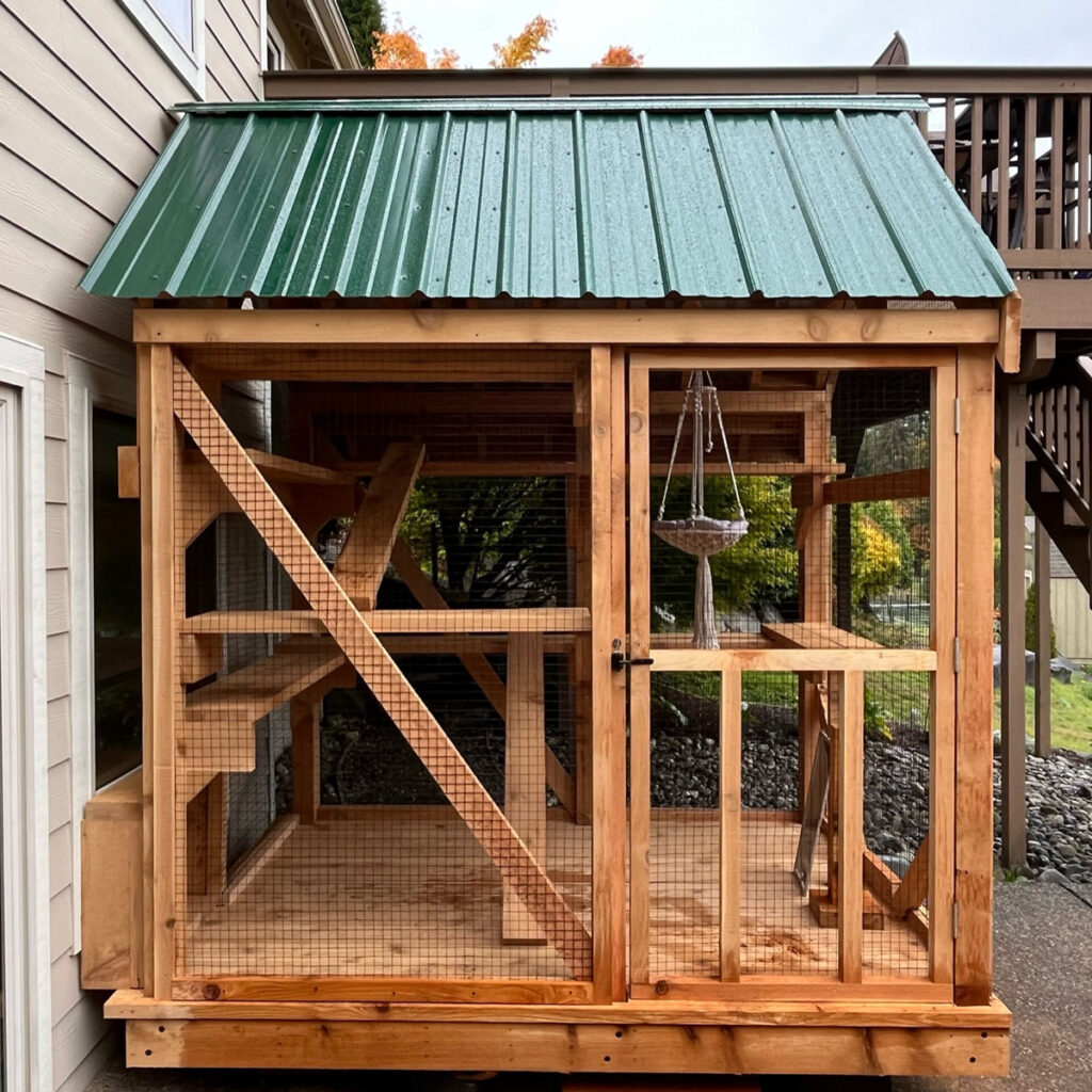 A compact yet spacious wooden catio with a green metal roof is securely attached to the side of a house. The enclosure features multiple wooden shelves, ramps, and a cozy macramé hanging hammock for feline relaxation. Sturdy wire mesh ensures safety while allowing fresh air and outdoor views. A small cat door connects the catio to the house, providing easy indoor-outdoor access.