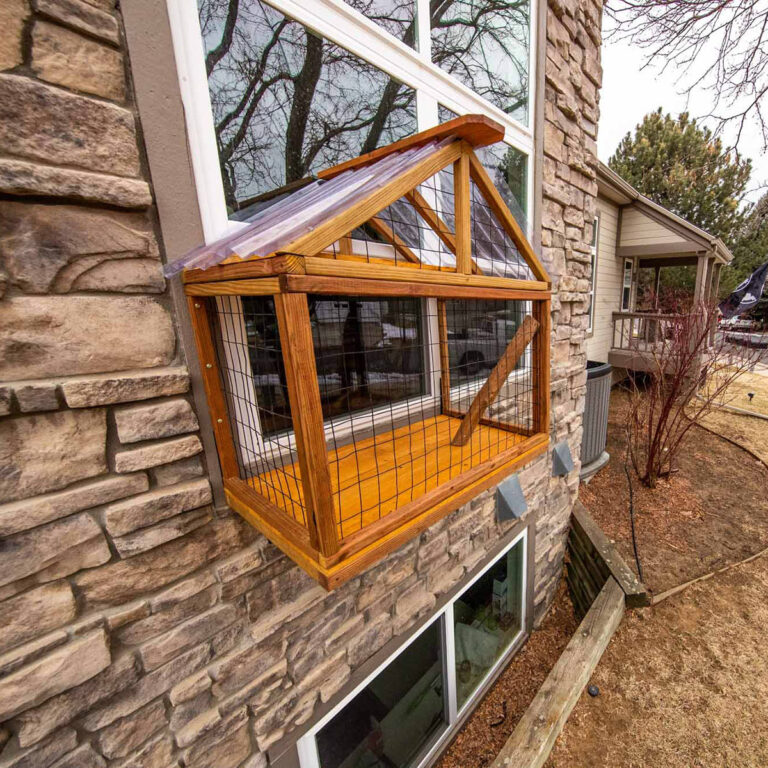 Elevated wooden catio with wire mesh and clear roof panels mounted on a stone exterior wall