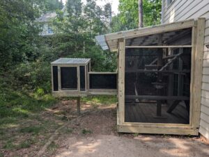 A custom-built catio attached to a house in Cary, North Carolina, featuring a large main enclosure with a slanted polycarbonate roof and a long elevated tunnel leading to a smaller secondary enclosure on a post, all surrounded by wire mesh for outdoor cat safety.