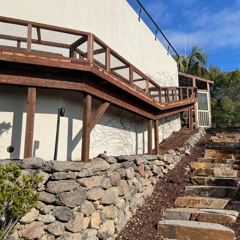Extended catio deck with mesh sides built along sloped wall above stone terrace and garden stairs.