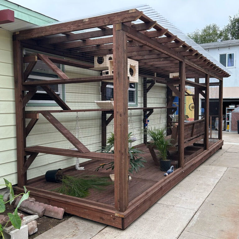 Angled view of long wooden catio with hanging toys, mounted shelves, greenery, and integrated cat houses.