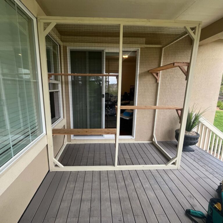 Front-facing view of a beige-framed small catio with wood shelves, installed on a deck outside a sliding glass door.