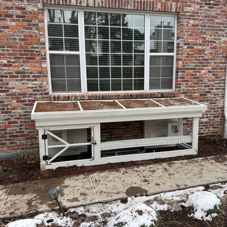 A low-profile white catio with a mesh enclosure and window access, positioned against a brick house and covered with a soil-coated roof.