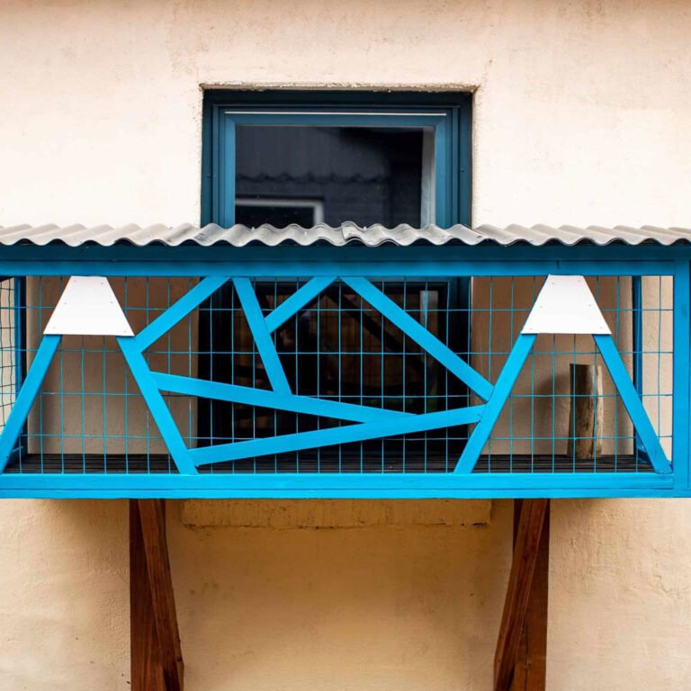 Front view of a small custom catio painted bright blue with wire mesh panels, angular wooden framing, and a corrugated roof, mounted below a window on a beige exterior wall.