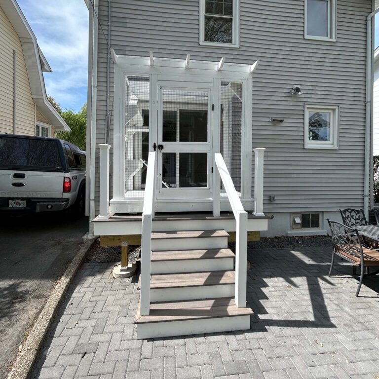 Front view of a small white catio enclosure connected to a home’s back door, featuring stairs, railings, a mesh screen door, and a paved patio area.