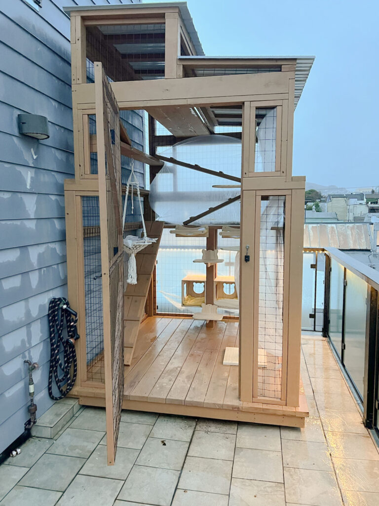 Outdoor catio with a high, enclosed vertical tunnel leading to a rooftop platform, surrounded by mesh panels and attached to the side of a house with direct window access.