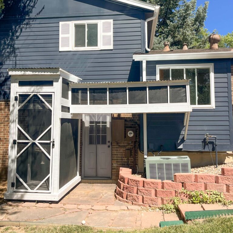 Custom white-framed catio tunnel and tower attached to a blue house, with an overhead mesh walkway connecting a tall vertical enclosure to a second-story window.