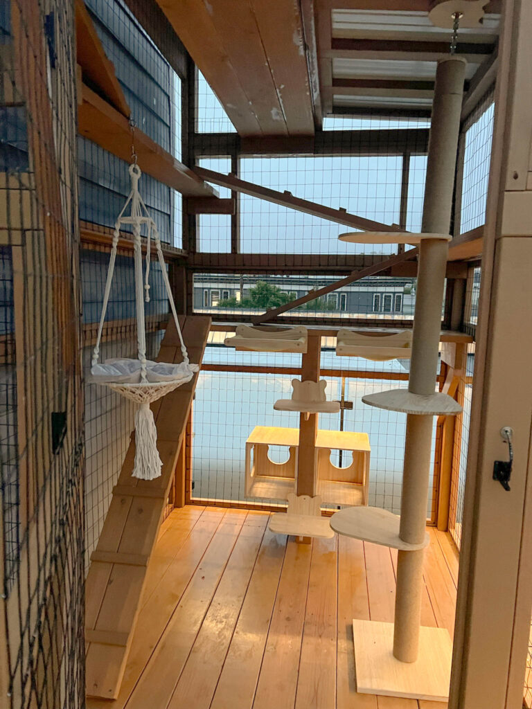 Custom-built outdoor catio featuring a wooden climbing ramp, sisal-wrapped vertical post with perches, modern cube-style shelves, and a macramé cat hammock under a wood-paneled roof.